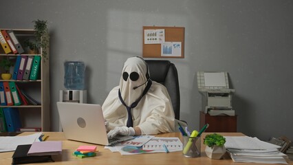 Halloween businessman ghost in white sheet sits at office desk works on laptop wearing headset and...