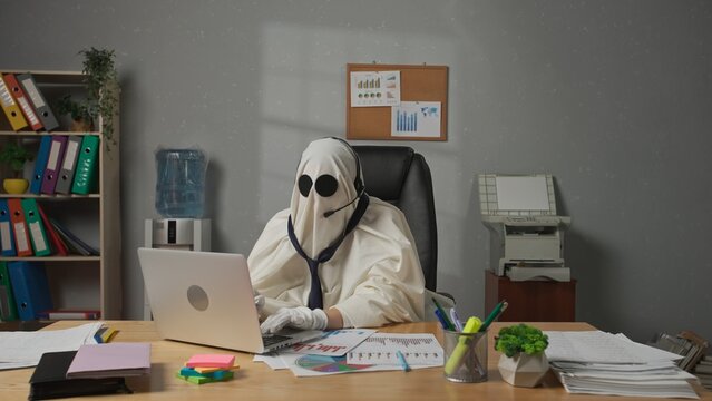 Halloween businessman ghost in white sheet sits at office desk works on laptop wearing headset and talking with clients. Happy halloween business concept.
