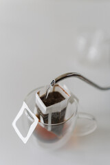 Boiling water pouring from kettle into drip coffee bag placed in glass cup, offering simple and...