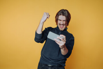 Young man celebrating a victory while playing a game on his smartphone against a bright yellow...