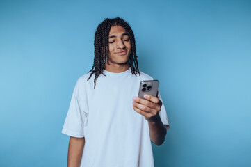 Fototapeta premium Young man with braided hair smiles while using smartphone against a blue background