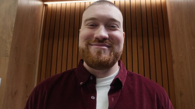 Low angle handheld portrait of cheerful gen Z male manager standing by multifunction printer smiling at camera in office corridor