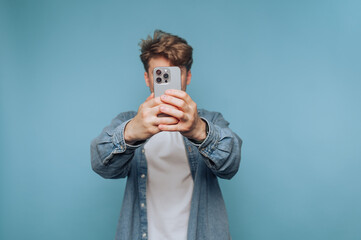 Young man takes selfie in front of blue wall, capturing a casual moment with a smartphone in hand...