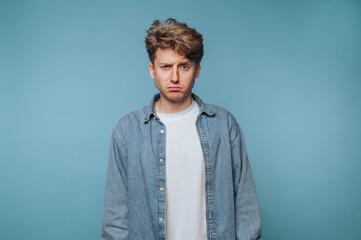 Young man with a serious expression standing against a light blue background in casual attire...