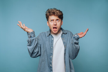 Young man with tousled hair expresses confusion with raised hands against a blue background