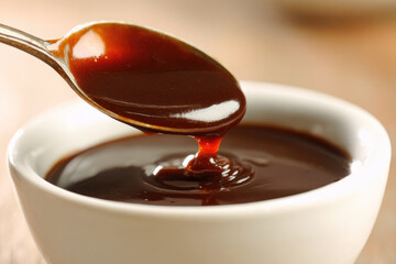 Thick brown sauce glossy texture metal spoon dipped in rich gravy white ceramic bowl close up savory appetizing comfort food smooth consistency warm lighting