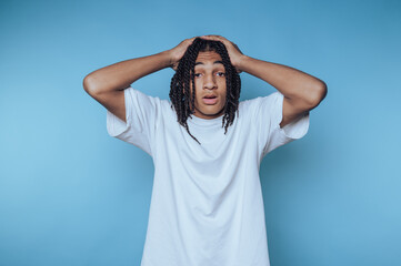 Young man expressing surprise against a blue background with hands on his head in a small indoor...