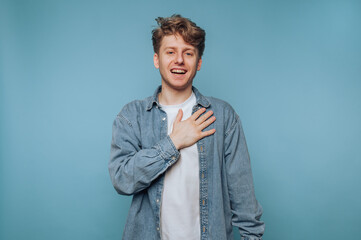 Young man expresses joy with hand on heart against blue backdrop in casual attire