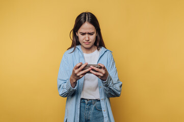 Woman with long dark hair looks frustrated while using her smartphone against a yellow background