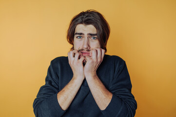 Nervous young man expresses anxiety against an orange background while biting his nails during a...