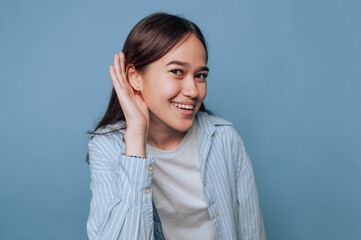 Young woman smiling and listening intently with a playful expression in front of a solid blue...