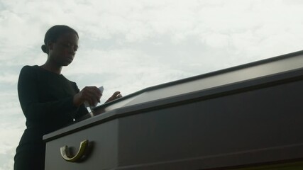 Medium shot of young Black widow in mourning dress approaching closed casket and grieving in silence at outdoor funeral