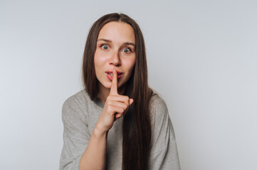 Woman with long hair gestures for silence while wearing a casual gray shirt against a plain...