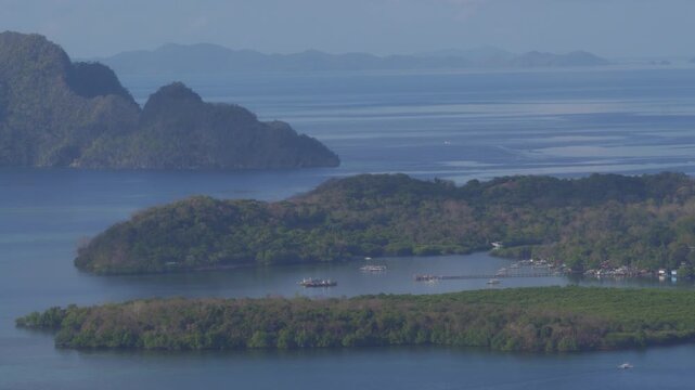 Drone glides above Mount Tapyas, unveiling mangrove-lined islets, a wooden pier and moored outriggers on glassy Coron Bay against hazy layered limestone peaks.