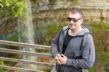 Fototapeta premium Smiling man in sunglasses and hoodie enjoying nature at a scenic waterfall viewpoint. Relaxed tourist on a wooden bridge in a canyon. Travel, adventure, lifestyle, and outdoor concept
