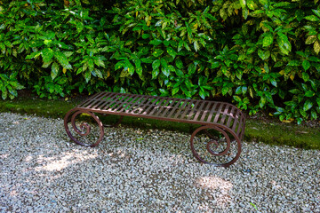 Ornate metal bench featuring scrollwork design is situated on gravel, surrounded by vibrant greenery and trees, providing a tranquil outdoor atmosphere. Ppark of villa Valsanzibio is a rare example 