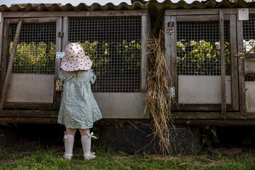 A young child in a light green dress and white hat with red polka dots stands near weathered wooden rabbit cages with wire mesh doors and hay.