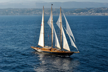 Fototapeta premium Elegant wooden sailing yacht with white sails cruising on open sea near coastal city with mountains in the background