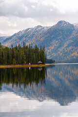 A serene lake reflects a shoreline with a small gazebo and pine trees, backed by snow-dusted mountains and autumn foliage.