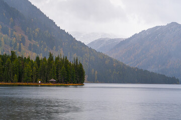 A serene mountain lake bordered by a dense forest, with mist-shrouded Altai mountains in the background under a cloudy sky.