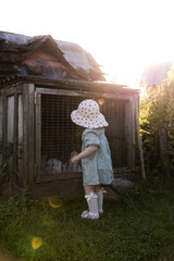A young child in a light blue dress and polka dot hat stands near a weathered wooden rabbit cage with wire mesh, surrounded by grass and plants.