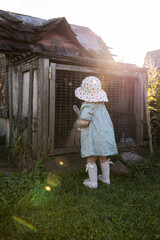 A young child in a light blue dress and white hat stands by a weathered wooden rabbit hutch, holding a gardening tool under warm sunlight with lens flares.
