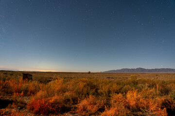 A vast desert landscape under a clear, starry night sky, with distant mountains and faint city...