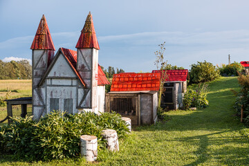 Outdoor scene with a whimsical castle like structure with red pointed roofs, rabbit cages nearby, green grass, plants, tree stumps, and open fields.