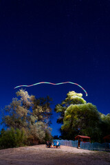 People sit by a campfire under a starry night sky, with long exposure light trails creating an...