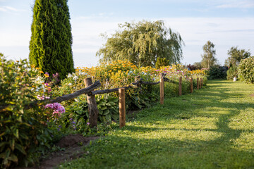 A grass pathway bordered by wooden posts and branches leads through a garden filled with colorful flowers, plants, an evergreen tree, and a weeping willow.