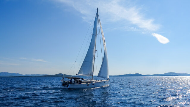 Sailing yacht gliding across the sea on a sunny day. View of the boat with full sails, clear sky, sparkling water and distant coastline in the background.