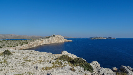 Rocky coastline with steep cliffs and deep blue sea, viewed from a high vantage point, with distant islands under a clear sky.