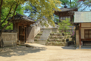 Traditional Korean hanok architecture with a wooden gate, tiled roof, stone steps, and spring greenery in a peaceful courtyard.