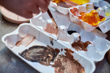 Close-up of hand mixing brown and orange paint in tray. Natural lighting highlights vibrant hues,...