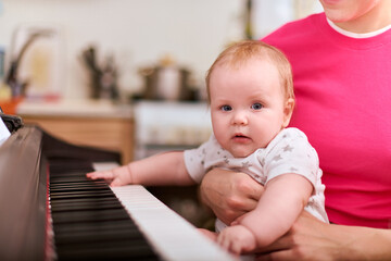 Fototapeta premium Baby girl, fair hair, held by young woman, explores piano in cozy room. Bright lighting highlights their joyful interaction and curious expression. Pink dominates