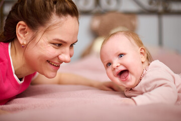 Young woman with brown hair and smiling baby girl on pink bed. Light-hearted interaction, creating...