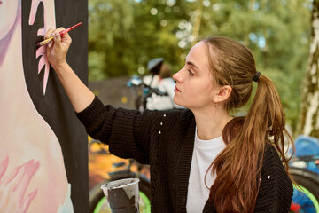 Young female with brown hair engaged in outdoor painting. Wearing black sweater, focused, creating...