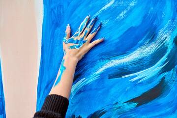 Adult female hand covered in vibrant blue paint creates expressive strokes on large canvas. Bold...