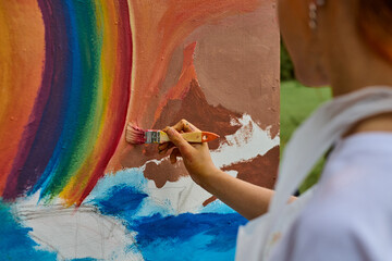 Young female artist with red hair paints vibrant mural in park. Canvas shows large rainbow, ocean...