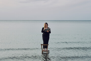 Young adult woman stands on chair in calm ocean, reading book under serene sky. Scene evokes...