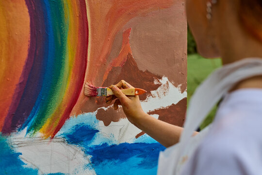 Young female artist with red hair paints vibrant mural in park. Canvas shows large rainbow, ocean landscape. Natural light enhances bright colors and creativity