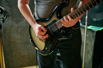 Fototapeta premium Musician playing an electric bass guitar at music rehearsal, practicing guitar technique in dimly lit setting, close up view guitarist hands