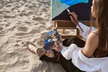 Young woman artist paints by sea, blending colors on palette. Her serene expression reflects...