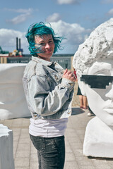 Young woman with teal hair and denim jacket stands near seaside area. Bright daylight illuminates...