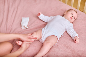 Young mother with brown hair, of caucasian descent, gently applies cream to baby girl lying on pink...