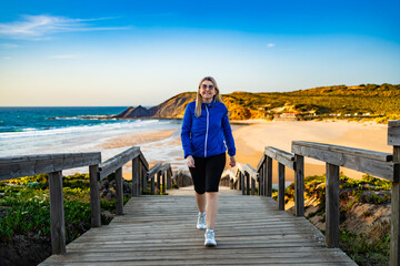 Beautiful mid adult woman walking up wooden stairs at viewpoint on Atlantic ocean and Amoreira beach in Portugal on spring evening. Front view