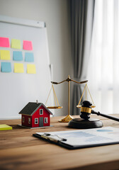 A wooden desk displays a model house, scales of justice, and a gavel, with colorful sticky notes on the wall in a bright office setting.