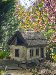 Small rustic miniature house with thatched roof in spring garden, surrounded by blooming pink flowers and greenery, captured in soft light.