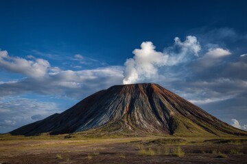 Dramatic landscape showcasing a volcano with smoke rising against a bright blue sky filled with clouds.