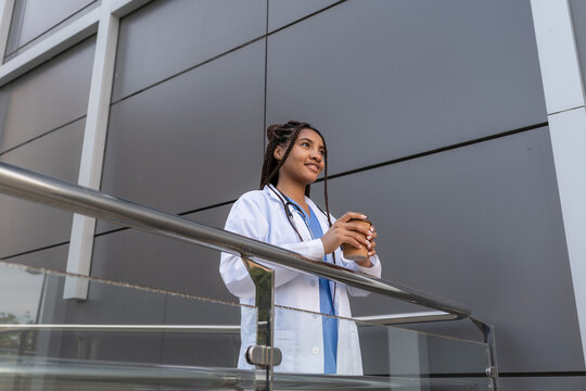 Smiling multiracial female doctor in white coat drinking coffee during break outside hospital